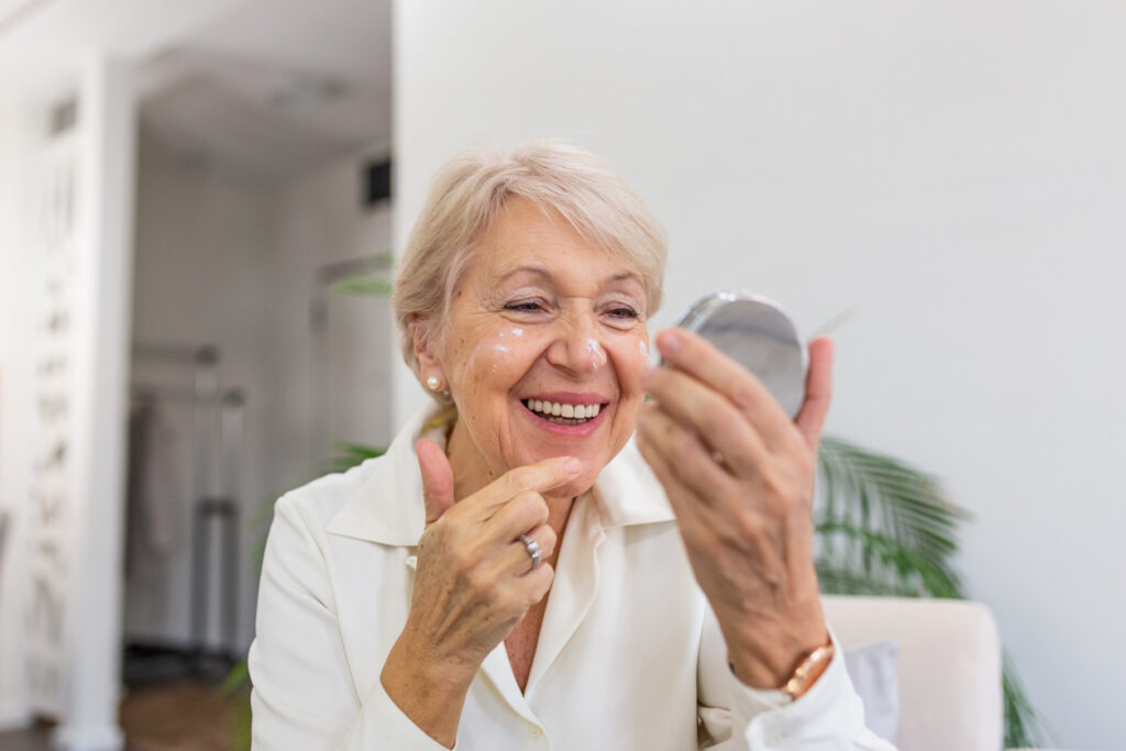 Charming, pretty, old woman touching her perfect soft face skin with fingers, smiling at camera over gray background, using day, night face cream, cosmetology procedures