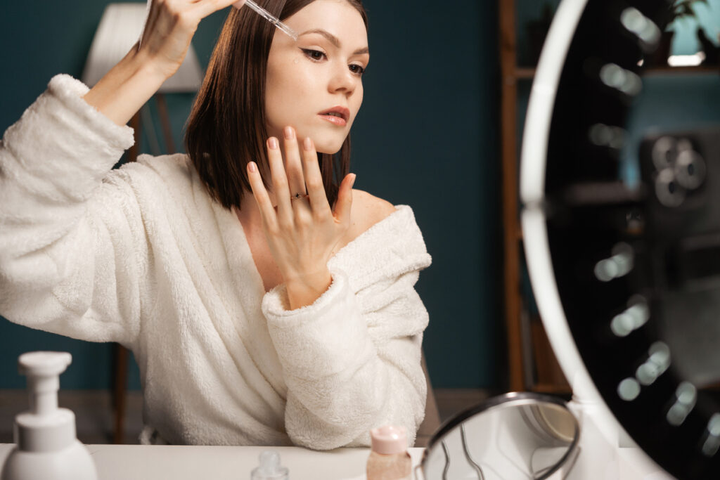 Beauty blogger applying face serum recording video on smartphone in holder with ring light