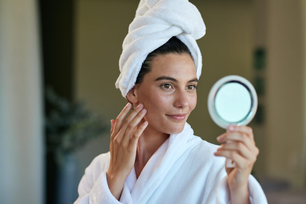 Cheerful woman gently checking blackheads and pores on her skin face while holding hand mirror. Young latin woman wearing a white bathrobe with towel turban enjoy a relaxing spa day. Hispanic girl feeling fresh after facial massage.